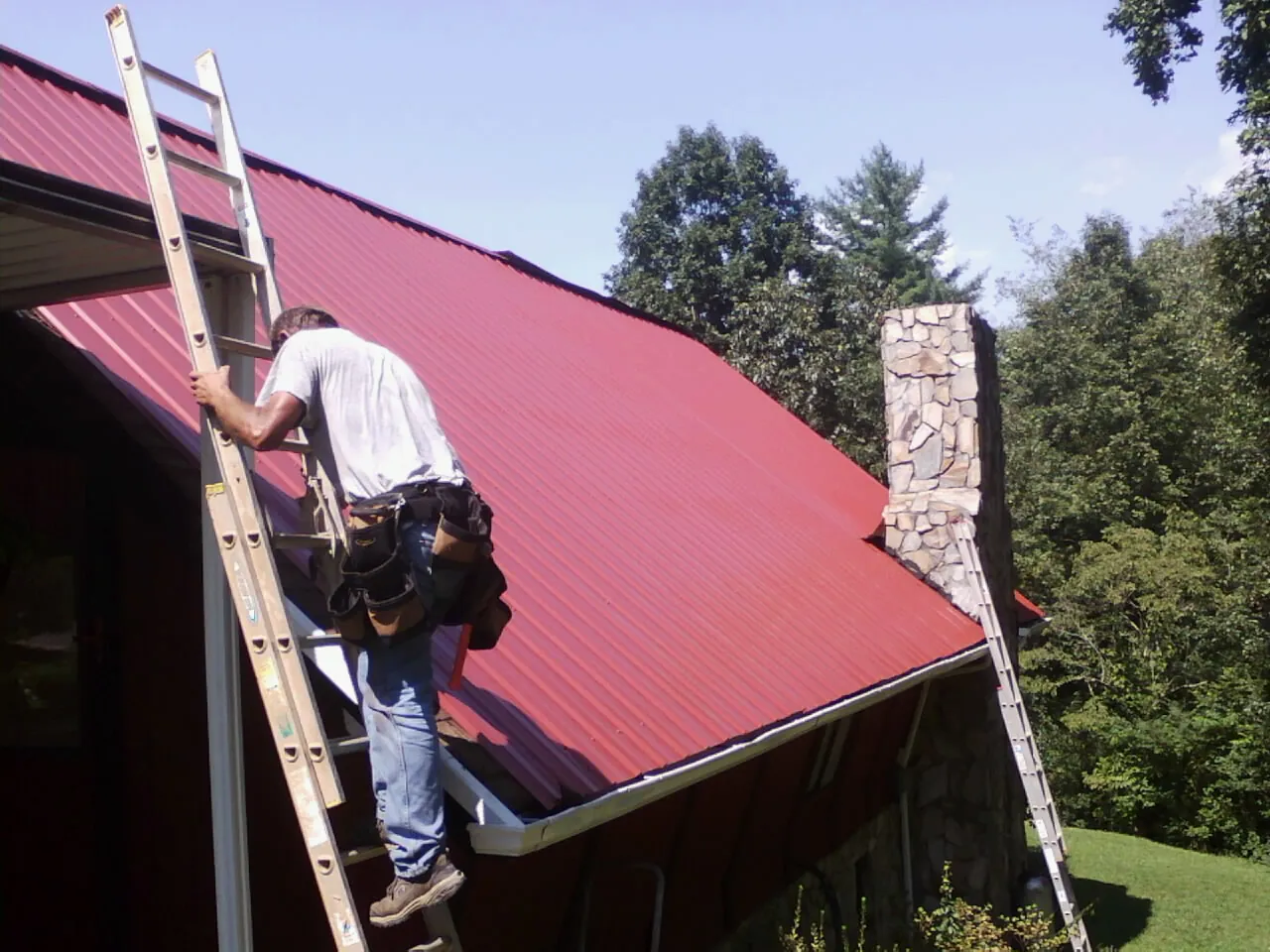 Roofer on a ladder beside a red metal roof and stone chimney — Soffit & Fascia Repair by Roofing Brothers in Plattsmouth